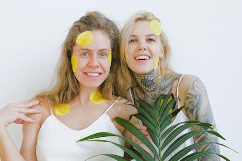 Happy women holding plant and looking at camera while standing and smiling while representing mask for skincare procedure and laughing