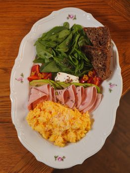 Healthy breakfast plate featuring scrambled eggs, ham, fresh spinach, tomatoes, and rye bread.