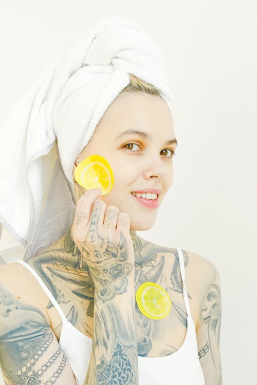 Cheerful young female with tattoo looking at camera while standing with slices of fresh citruses patches covering face while enjoying skincare