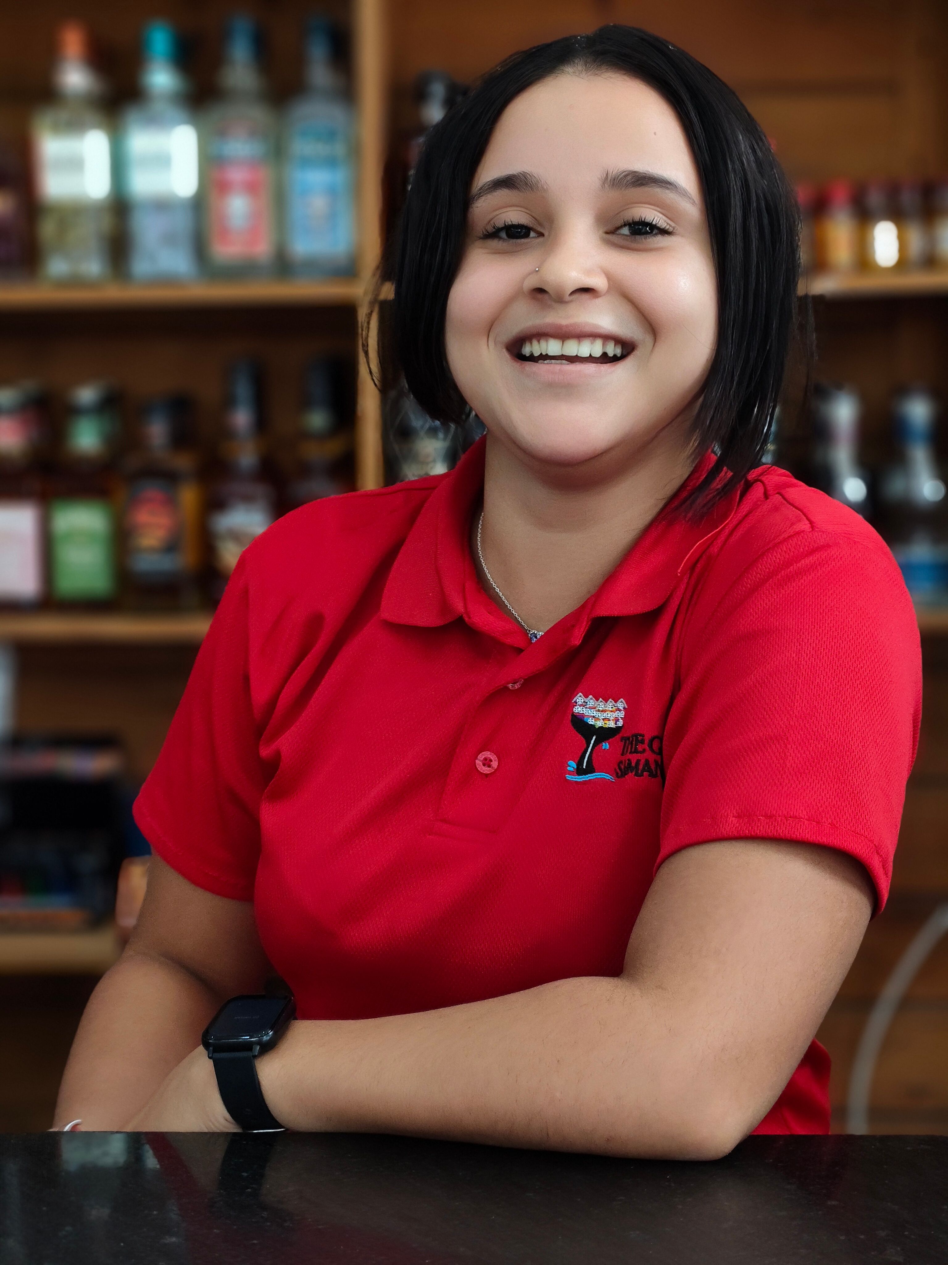 Cheerful woman in a red polo smiling inside a bar with bottles in the background.