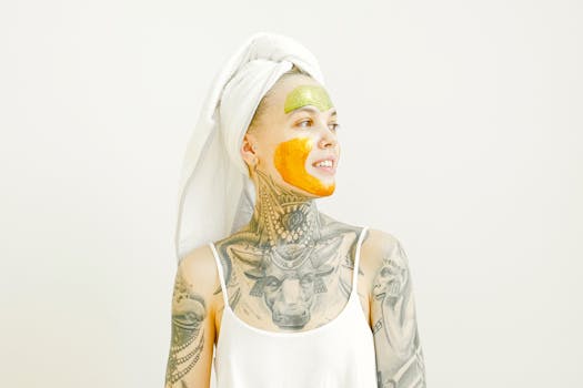Tattooed woman with orange and green face mask, towel wrapped around hair, relaxing indoors.