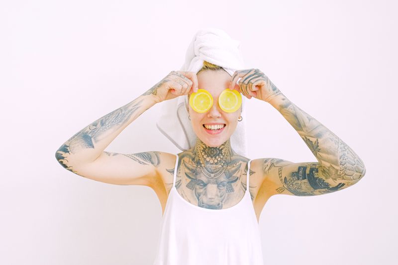 Cheerful tattooed woman with lemon slices and towel in studio, showcasing wellness and humor.