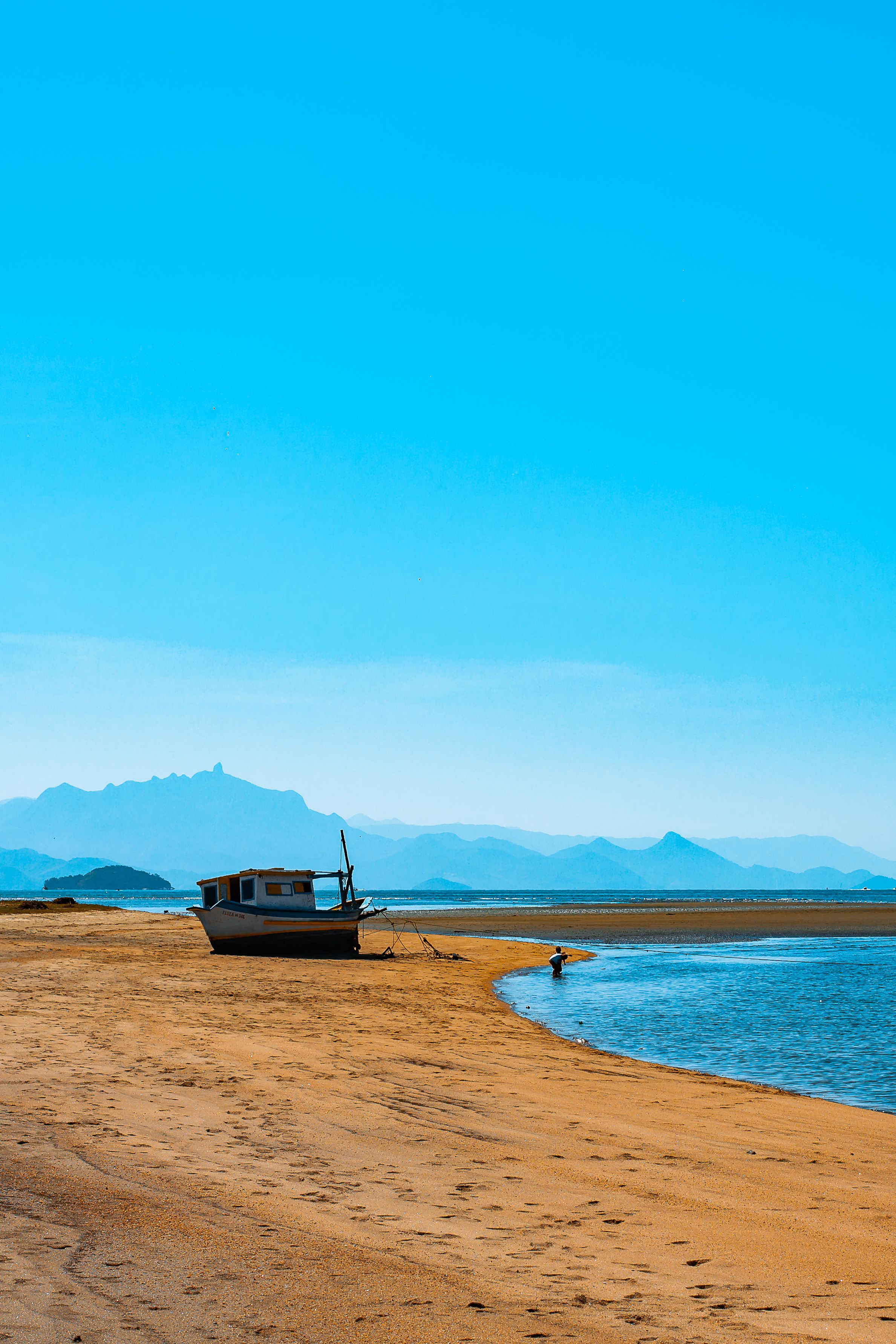 Serene beach with a boat and a person walking, under a clear blue sky.