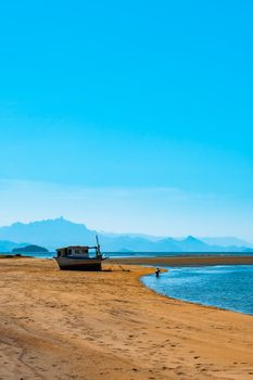 Serene beach with a boat and a person walking, under a clear blue sky.