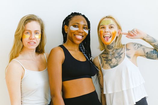 Excited young women smiling and looking at camera while having fun during weekend having beauty procedure with patches and mask