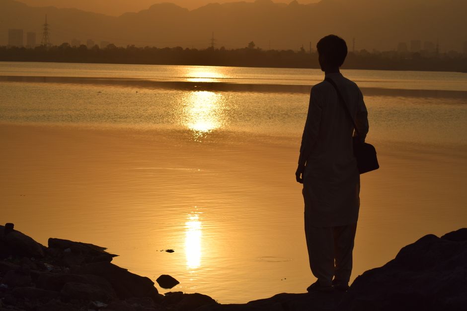 A man silhouetted against a golden sunset over a tranquil lake in Islamabad, Pakistan.
