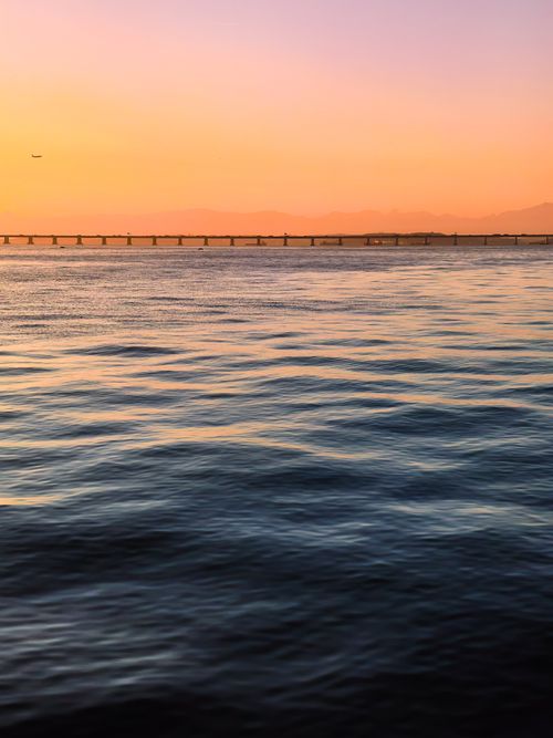 Free Serene ocean waves at sunset with a distant bridge horizon and gentle pastel sky. Stock Photo