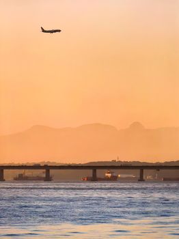 Airplane flying over bridge at sunset, with mountains in backdrop and boats on water beneath.