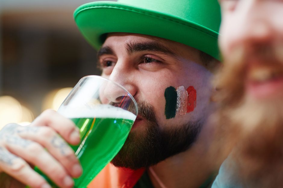 Close-up of a man enjoying green beer with an Irish flag painted on his cheek during St. Patrick's Day.
