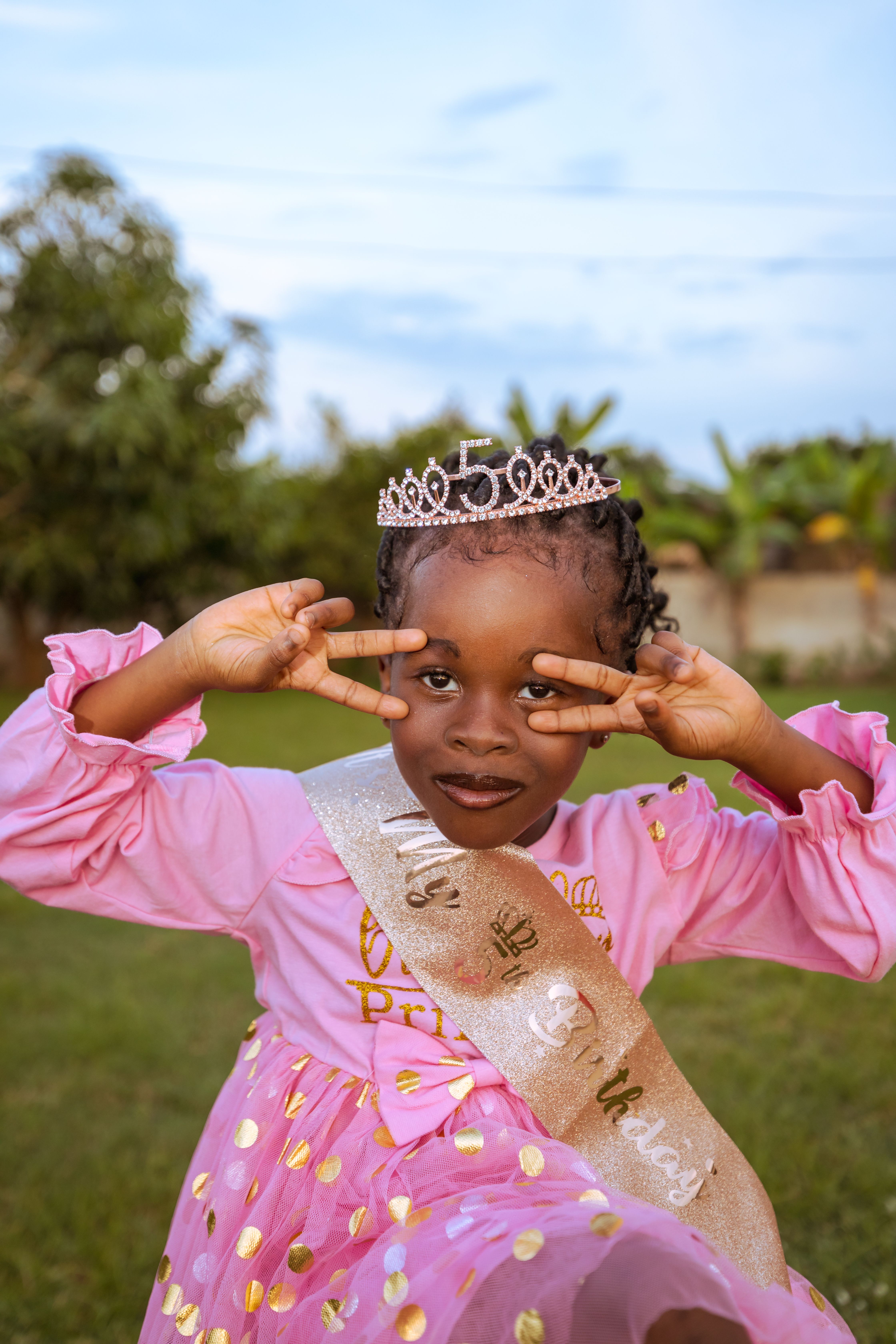 Young girl celebrating her fifth birthday with a tiara and playful pose in an outdoor setting.