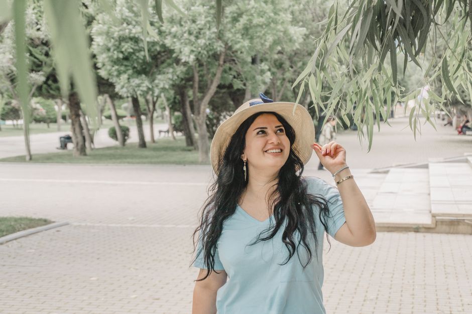 Happy woman wearing a hat and casual outfit smiles under leafy branches in a park in Baku, Azerbaijan.