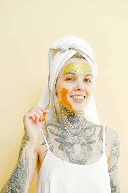 Young female with tattooed body smiling and looking at camera while removing peel off golden mask on yellow background