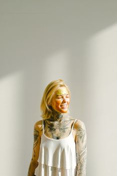 Portrait of a blond woman with tattoos wearing a face mask in bright sunlight. Indoor setting with soft shadows.
