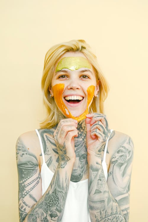 Young cheerful female with tattooed body and blond hair removing peel off golden mask from face looking at camera