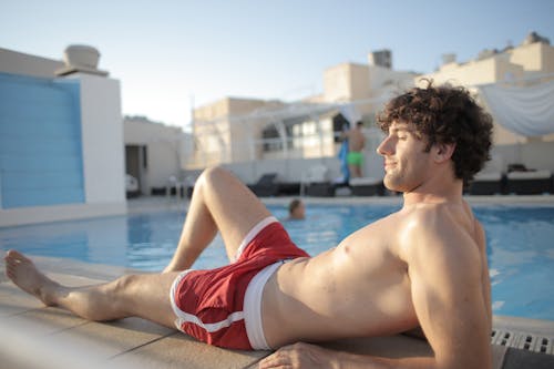Man in Red Shorts Sitting on Concrete Floor Near Swimming Pool