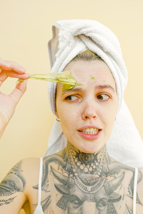 Young tattooed female with white towel on head gently removing peel off golden mask from face looking away
