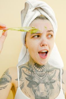 Young female with tattooed body in towel on head feeling uncomfortable while removing peel off mask