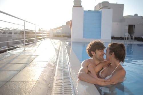 Cheerful couple in swimsuits cuddling in swimming pool with blue water near fenced embankment and looking at each other in sunlight