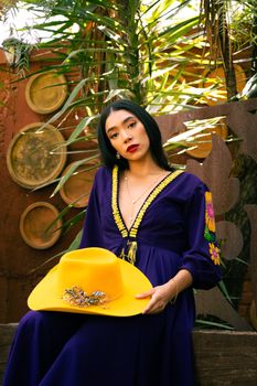 Woman in colorful dress poses gracefully with a vibrant yellow hat in a lush garden setting.