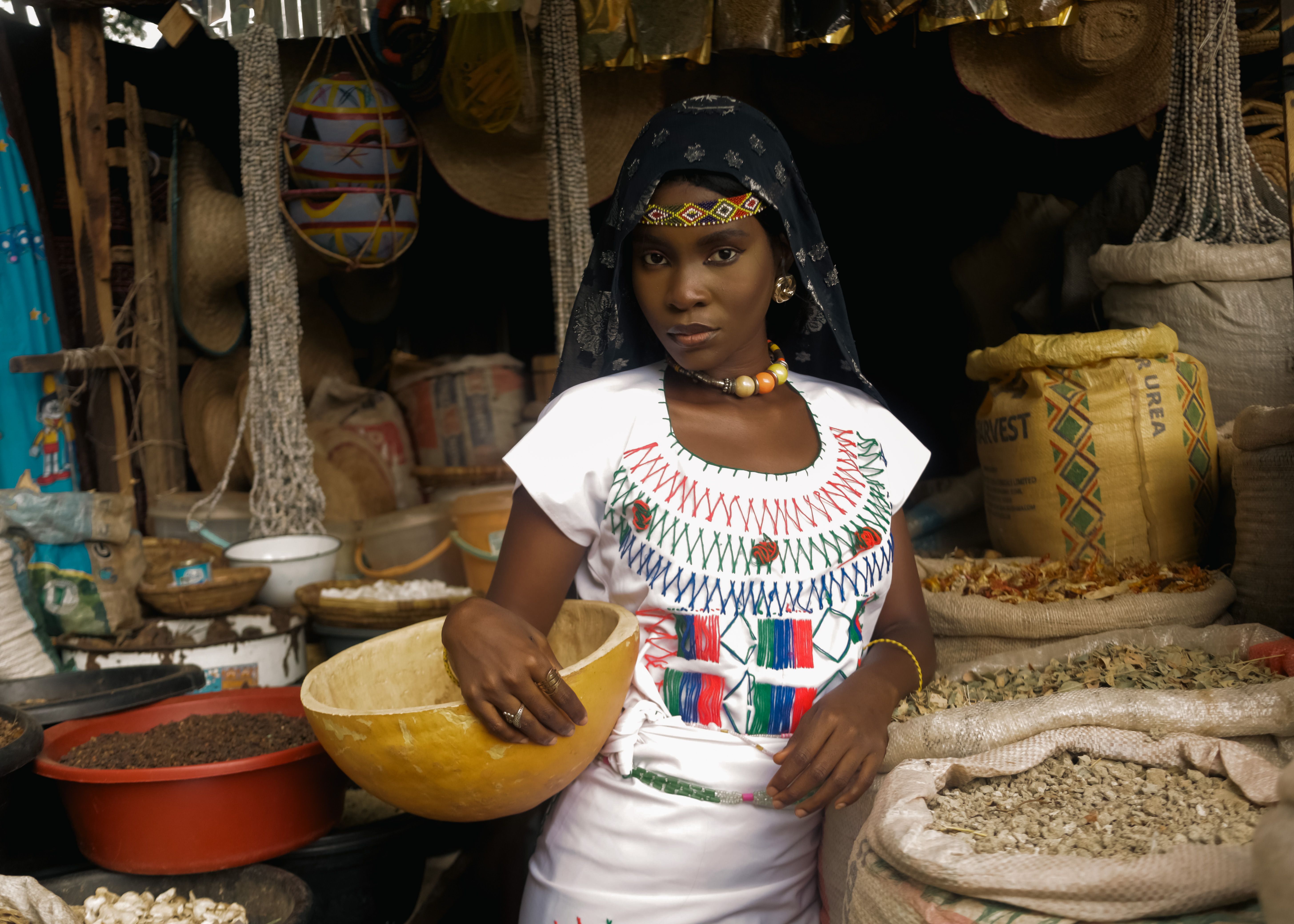 A female vendor in traditional attire at an African market stall filled with colorful textiles and grains.