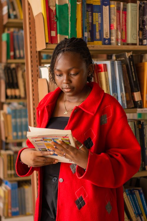 Free A young woman in a red coat reads a book in a library filled with colorful books. Stock Photo