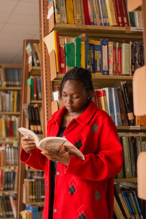 Free A young woman in a red coat reading a book in a library setting. Stock Photo