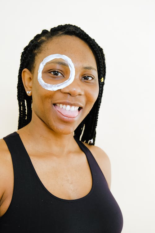 Happy African American lady in black wear with lotion circle around one eye showing tongue and looking at camera while standing on white background