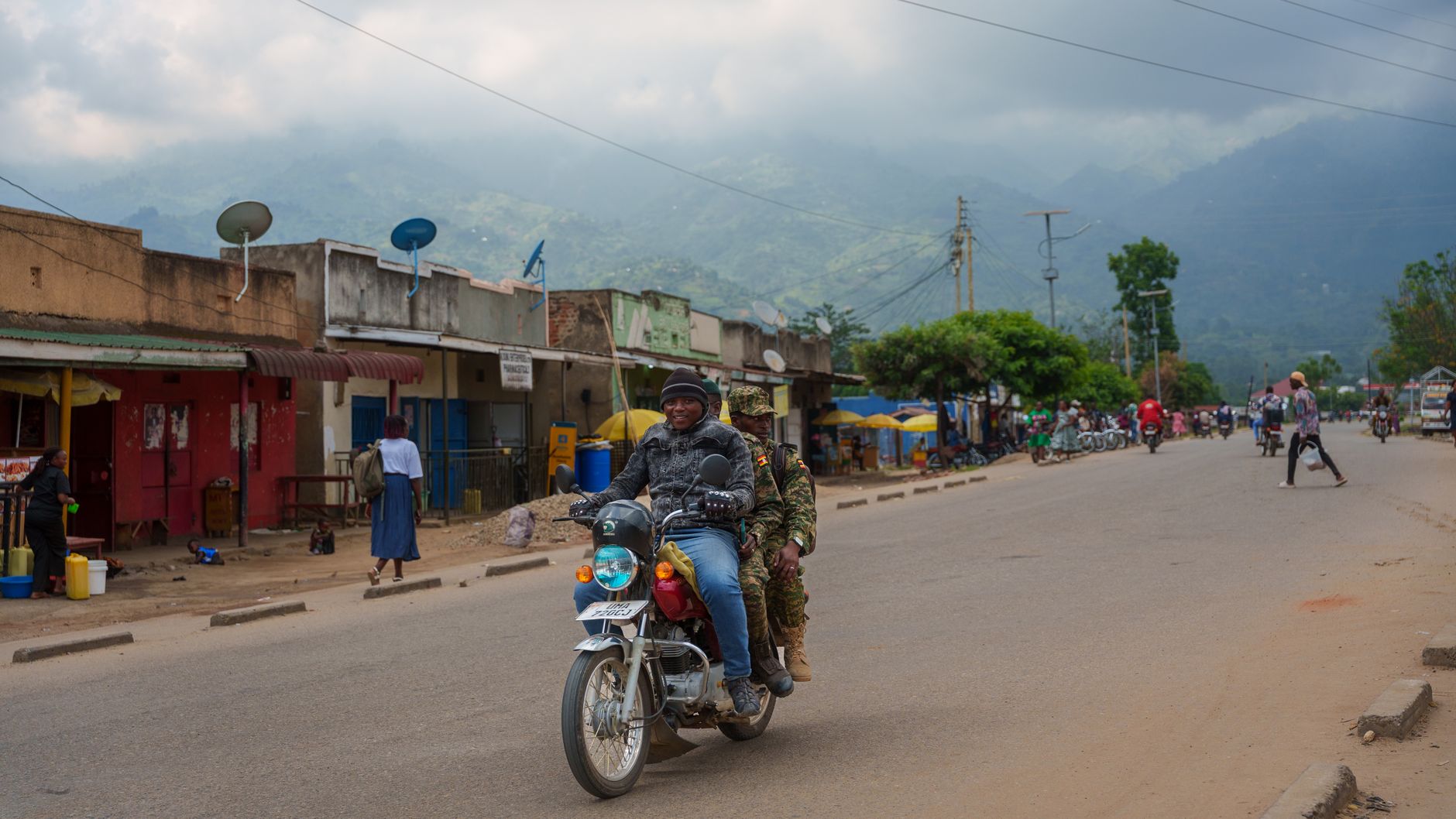 Motorcyclists ride down a bustling street in Uganda with scenic mountain backdrop.