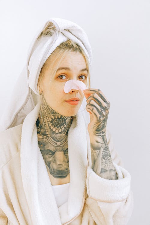 Free Young tattooed female in terry robe and towel taking off cosmetic nose mask while standing near white wall and looking at camera Stock Photo