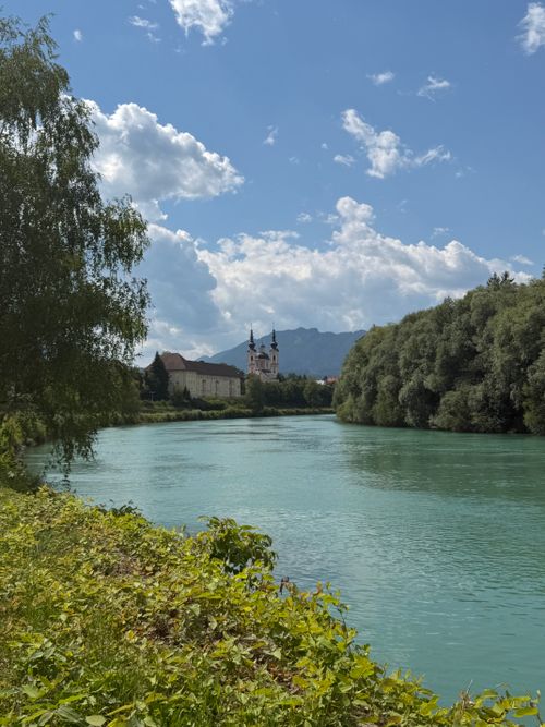 Free Idyllic scenery of the Drava River with lush green trees and distant church in Villach, Austria. Stock Photo