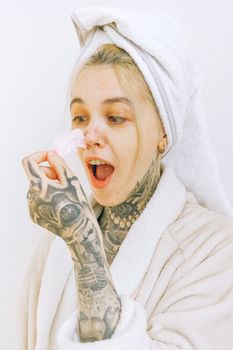Tattooed woman enjoying a skincare routine with a pore strip in a cozy robe and towel wrap.