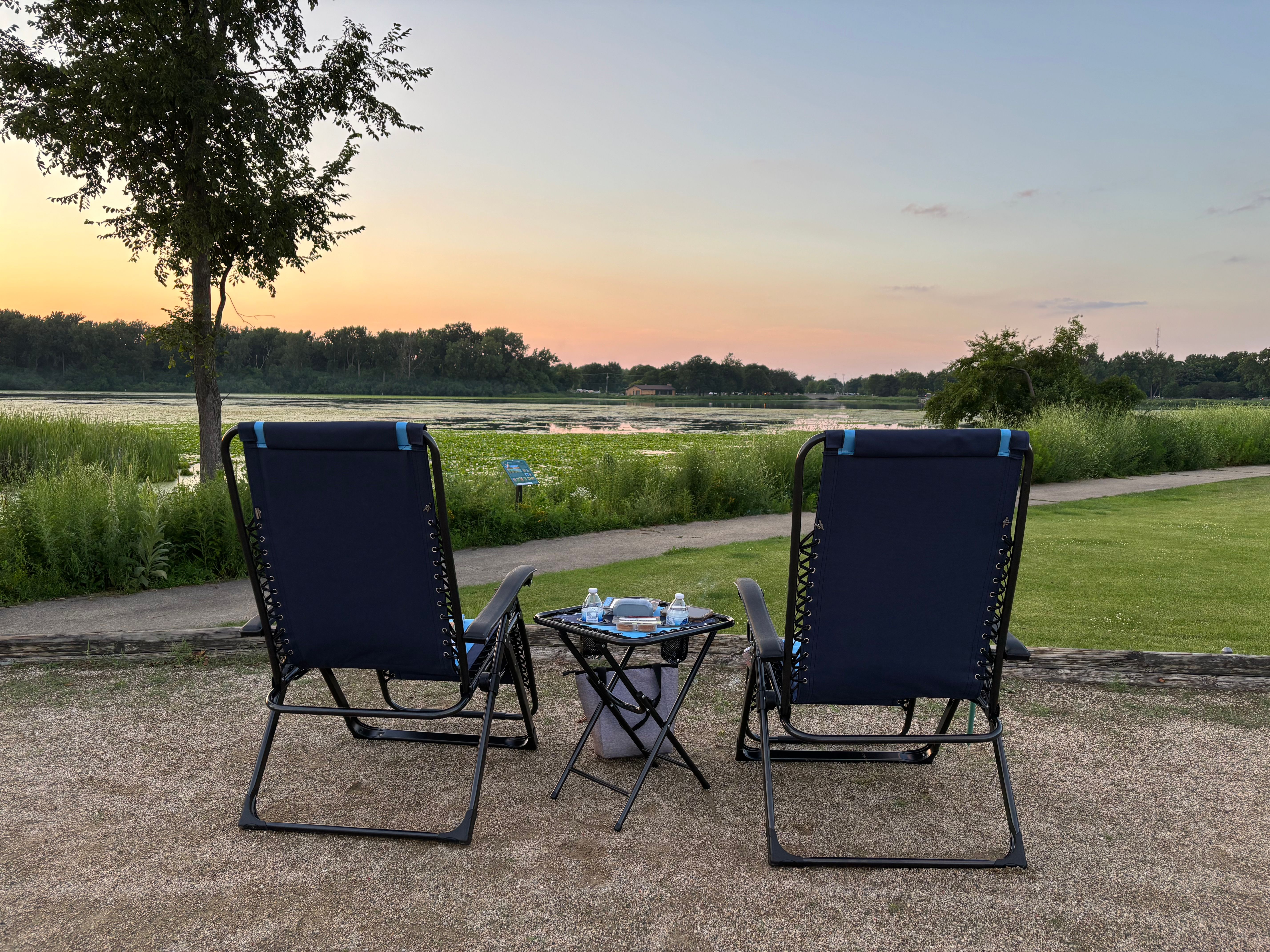 Relaxing lakeside sunset view with two chairs and a table set for leisure by the water.