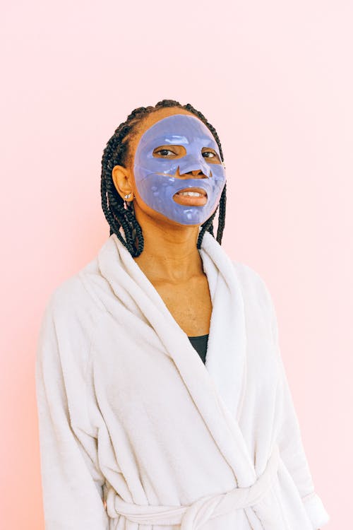 African American female in white terry robe with collagen mask applied on face standing on pale pink background and looking at camera