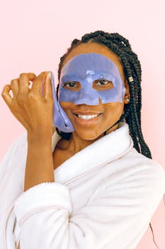 Smiling woman in bathrobe applying a purple face mask on a pink background.
