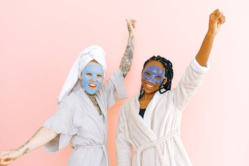 Free Happy diverse women in robes and blue collagen masks applied on face standing with raising hands near pale pink wall and looking at camera Stock Photo