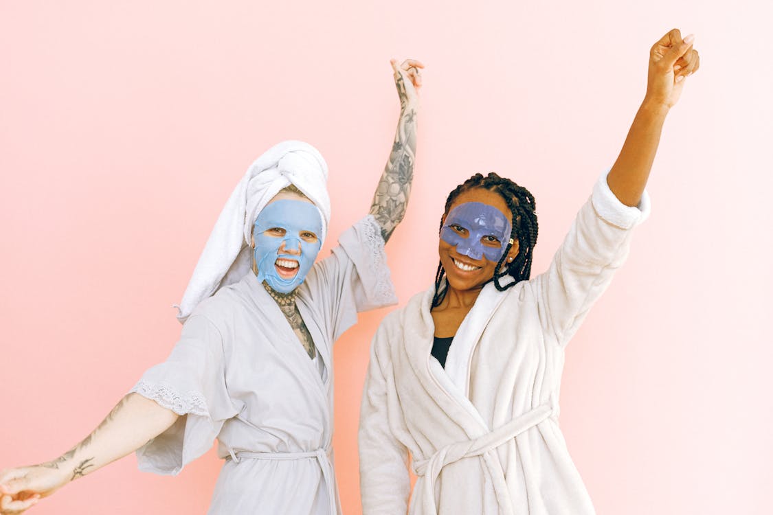 Two women enjoying a facial mask while wearing robes and smiling in front of a pink background