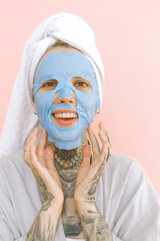 Content tattooed lady in peignoir and bath towel on head applying blue removable face mask while laughing and looking at camera