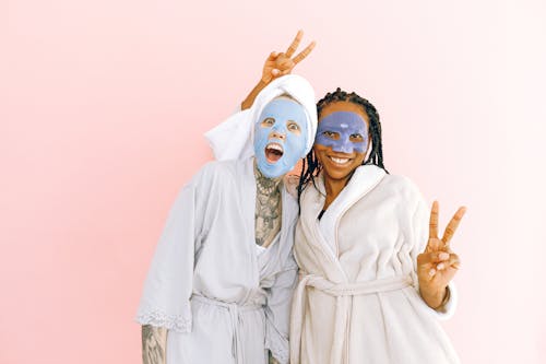 Free Cheerful young diverse ladies in robes and cosmetic masks applied on face standing on pale pink background and showing peace gesture while looking at camera Stock Photo