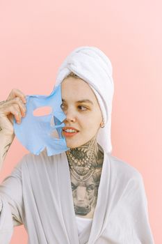 Surprised young tattooed female in dressing gown and terry bath towel taking off blue collagen face mask while standing on pink background and looking away