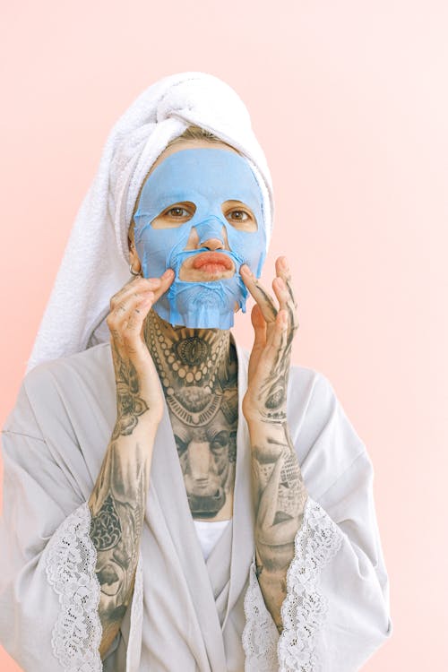 Free Young tattooed lady in peignoir and terry bath towel on head applying removable face mask while doing grimace with lips and looking at camera Stock Photo