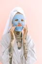 Young tattooed lady in peignoir and terry bath towel on head applying removable face mask while doing grimace with lips and looking at camera