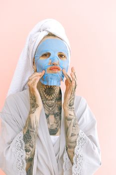 Young tattooed lady in peignoir and terry bath towel on head applying removable face mask while doing grimace with lips and looking at camera
