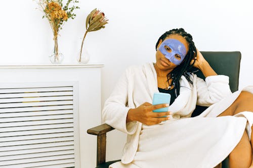 Thoughtful African American female in terry robe and blue collagen mask applied on face sitting on chair in spa salon and browsing internet on cellphone