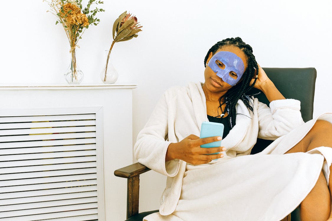 African American woman in bathrobe and cosmetic mask watching smartphone