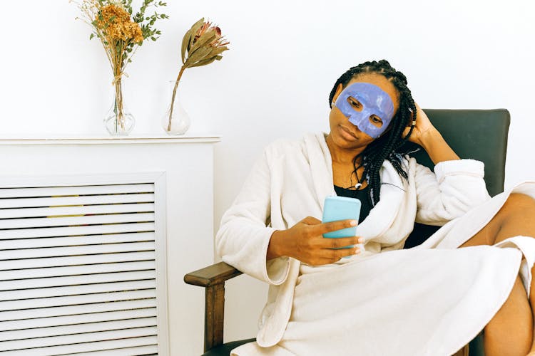 African American Woman In Bathrobe And Cosmetic Mask Watching Smartphone