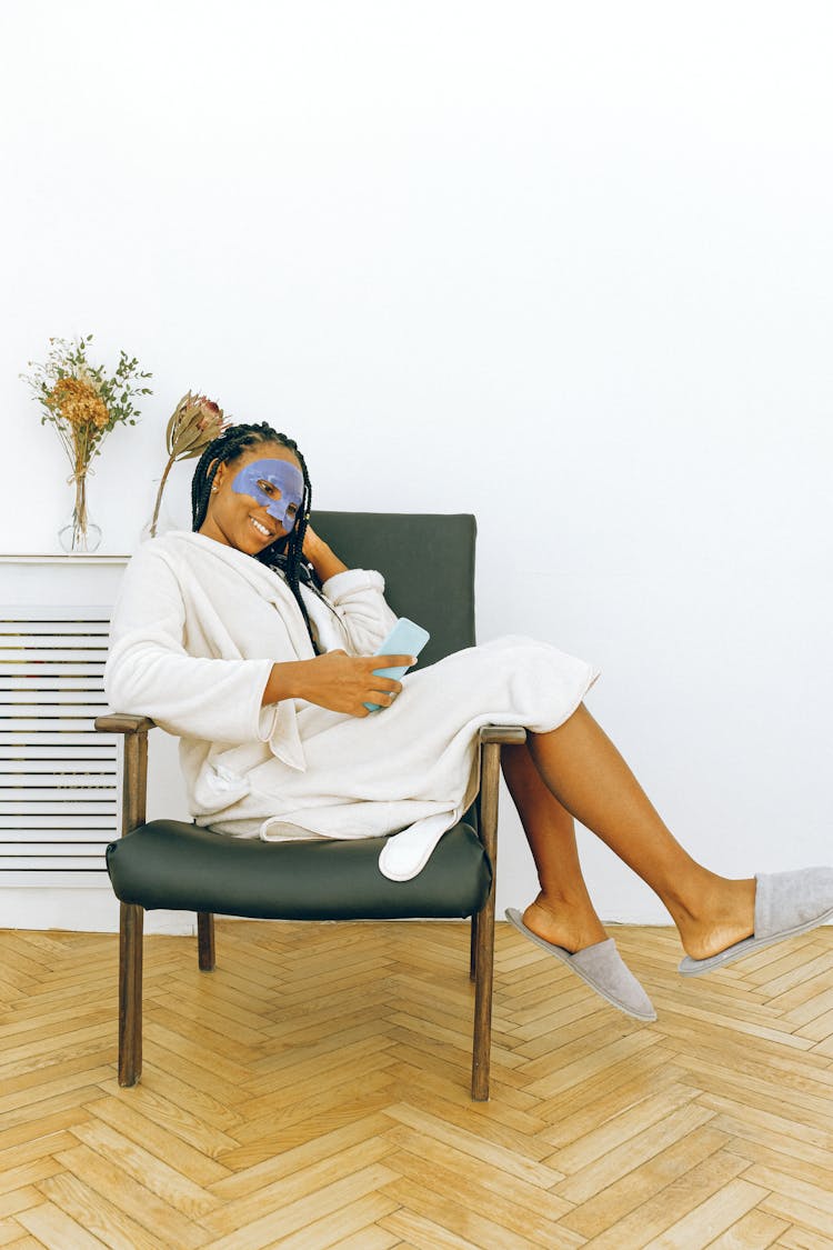 Young Cheerful Black Woman Relaxing With Smartphone During Facial Treatment At Home