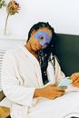 Young satisfied African American female with braided long curly hairstyle wearing white bathrobe sitting across chair while using cellphone during home cosmetic procedure for face skin on weekend