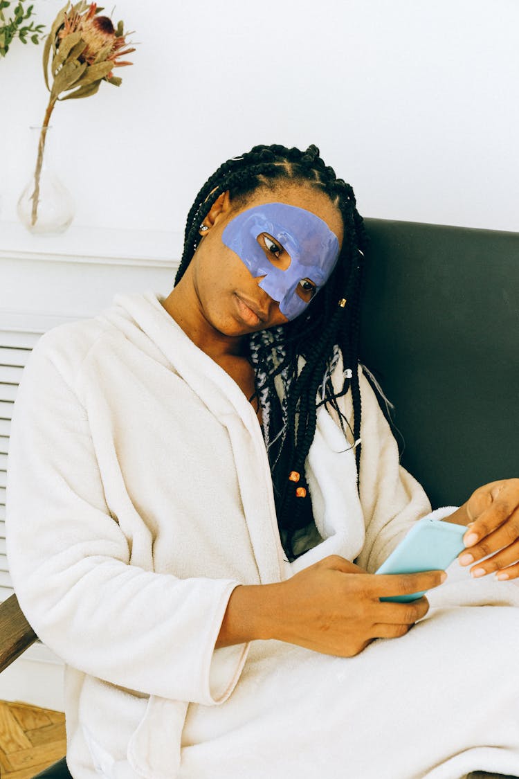 Relaxing Black Woman With Blue Facial Treatment Mask On Face With Smartphone