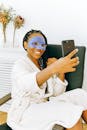 Cheerful African American female with braided long curly hairstyle wearing white bathrobe sitting across chair while making selfie with smile during home cosmetic procedure for face skin on free time