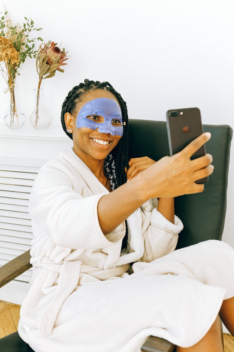 Young Black Woman In Blue Facial Treatment Mask Using Smartphone At Home